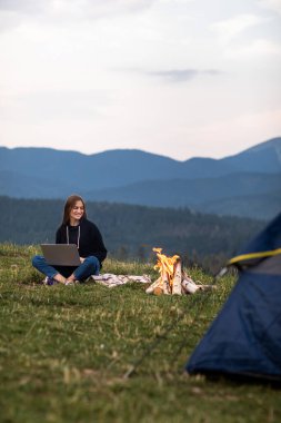 Genç bayan serbest yazar akşamları dağlarda dizüstü bilgisayarda çalışıyor. Kamp ateşi ve çadırın yanında oturan turist kız. Boşluğu kopyala.
