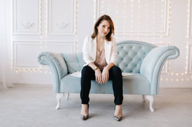 Beyaz ceketli, siyah pantolonlu ve yüksek topuklu ayakkabılı çekici genç bir kadın mavi klasik bir kanepede oturuyor. Işıklı beyaz arkaplan.
