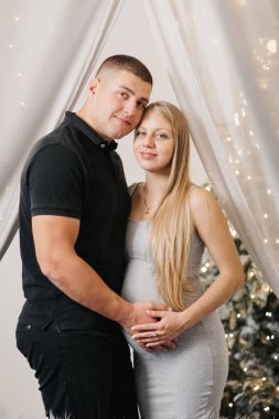 Evde Noel ağacının yanında sevimli bir çift, şefkatli koca güzel hamile eşe sarılıyor, genç aile mutluluğun tadını çıkarıyor, kış tatili konsepti.