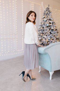 Beyaz kazak, etek ve yüksek topuklu ayakkabı giyen genç, çekici bir kadın Noel ağacının yanındaki kanepenin yanında.