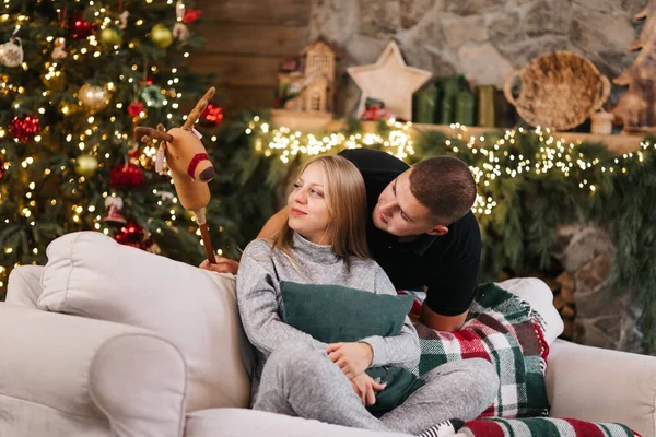 Adorable marriage couple sit near Christmas tree and fireplace at home, caring husband hug beautiful pregnant wife, young family enjoy happiness, winter holidays concept.