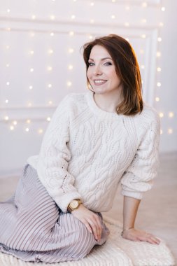 Beyaz bir kazak, etek ve topuklu ayakkabıyla beyaz bir duvar ve Noel çelengi giymiş güzel genç bir kadın..