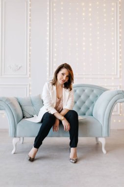 Beyaz ceketli, siyah pantolonlu ve yüksek topuklu ayakkabılı çekici genç bir kadın mavi klasik bir kanepede oturuyor. Işıklı beyaz arkaplan.