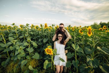 Güneş gözlüklü güzel bir çift ayçiçeği tarlasında eğleniyor. Aşık bir adam ve bir kadın bir tarlada ayçiçekleriyle yürür, bir erkek bir kadına sarılır. seçici odak.