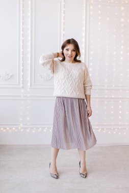 Beyaz bir kazak, etek ve topuklu ayakkabıyla beyaz bir duvar ve Noel çelengi giymiş güzel genç bir kadın.