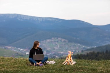 Genç bayan serbest yazar akşamları dağlarda dizüstü bilgisayarda çalışıyor. Kamp ateşinin yanında oturan turist kız eğleniyor. Boşluğu kopyala