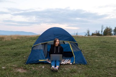 Dağlarda dizüstü bilgisayarı olan kız. Turist kadın doğada bir dizüstü bilgisayar kullanıyor, bir dağ manzarasının arka planına karşı bir yolculukta çadırın yakınındaki bir bilgisayarda daktilo yazan bir serbest yazar.