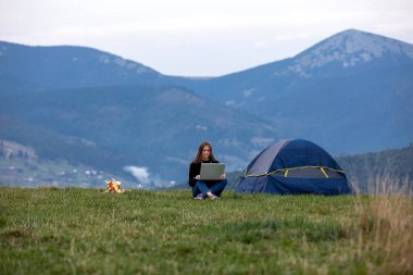 Genç bayan serbest yazar akşamları dağlarda dizüstü bilgisayarda çalışıyor. Kamp ateşi ve çadırın yanında oturan turist kız. Boşluğu kopyala.
