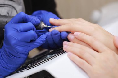 Güzellik salonundaki donanım manikürünü yakından çek. Koruyucu eldivenli manikürcü kadın parmaklarına manikür yapmak için elektrikli tırnak matkabı uyguluyor.