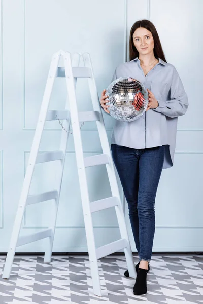 beautiful young woman with a disco ball in her hands. girl with long hair in blue clothes near stepladder on blue wall background.