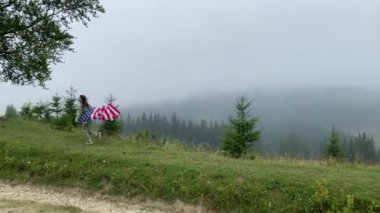 Mutlu küçük kız bulutlu dağlarda koşuyor ve kaygısız atlıyor. Amerikan bayrağı taşıyor. seçici odak.