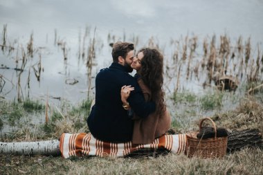 Gölün kenarında battaniyenin üzerinde oturan iki Kafkas aşığı. Genç çift, sonbahar günü dışarıda kucaklaşıyor. Sakallı bir adam ve birbirine aşık kıvırcık bir kadın. Sevgililer Günü. Aşk ve aile kavramı. arka plan.