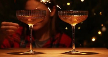 İki kadeh şampanya ve altın ışıklı karanlık bir arka plan ve arka planda yeni yıl ışıltısıyla odaklanmış bir kadın portresi.