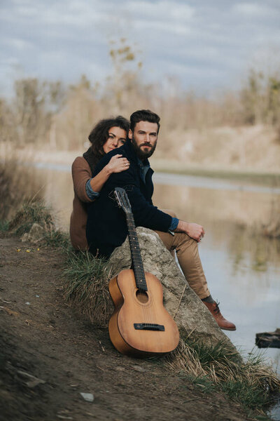 caucasian man is playing guitar with woman by the lake. Young couple is hugging on autumn day outdoors. A bearded man and curly woman in love. Valentine's Day. Concept of love and family