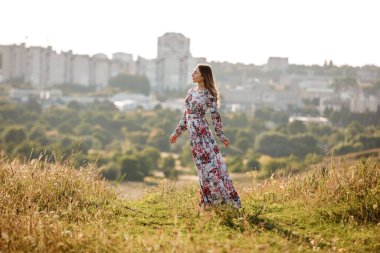Çiçek desenli giyinmiş güzel bir kadın gün batımında tarlada yürüyor. Uzun saçlı şık romantik kız şehir arka planında iyi vakit geçiriyor. Boşluğu kopyala.