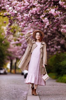 Rüzgarda uçan uzun saçlı ve çiçek açan bir bahçede kahverengi şapkalı güzel esmer kadın. Kalın bir alan derinliği. bahar günü.