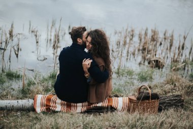 Gölün kenarında battaniyenin üzerinde oturan iki Kafkas aşığı. Genç çift, sonbahar günü dışarıda kucaklaşıyor. Sakallı bir adam ve birbirine aşık kıvırcık bir kadın. Sevgililer Günü. Aşk ve aile kavramı. arka plan.