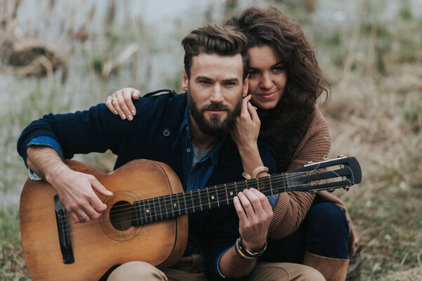 caucasian man is playing guitar with woman by the lake. Young couple is hugging on autumn day outdoors. A bearded man and curly woman in love. Valentine's Day. Concept of love and family