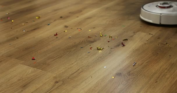 Vacuum cleaner robot putih otomatis dengan sikat membersihkan confetti dari lantai kayu setelah Natal, pesta ulang tahun. pembersihan otomatis apartemen pada waktu tertentu. — Stok Video