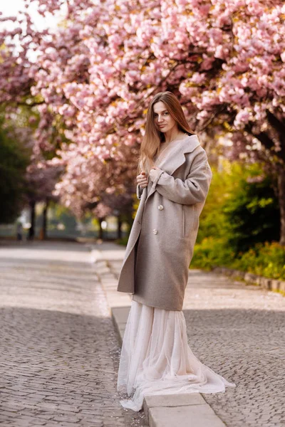 女人的日子 美丽的年轻女子穿着华丽的长裙 接近樱花盛开 在春天公园的背景上 在盛开的樱花旁 一个时髦的女孩正在绽放 与自然和谐相处概念 — 图库照片