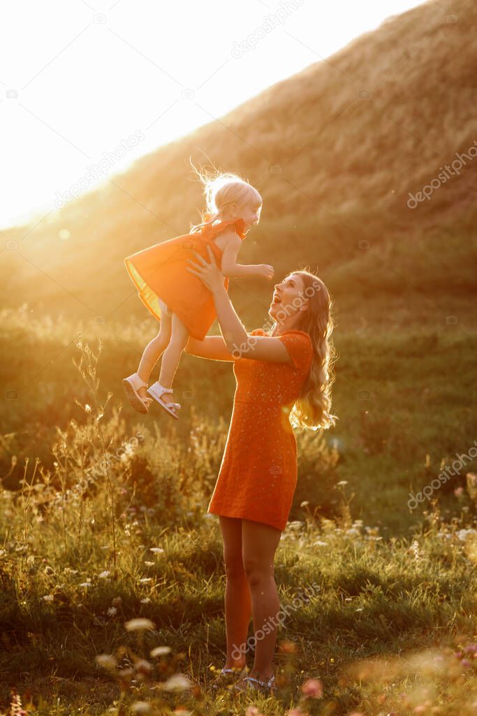 Portrait of a mother holds, throws up and spins the daughter on hands on nature on summer day vacation. Mom and girl playing in the park at the sunset time. Concept of friendly family. Close Up.