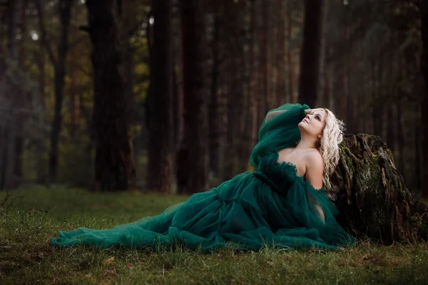 Uzun yeşil elbiseli güzel sarışın bir kadın ve ormanda kafasında bir diyabet. Yaşlı kütüğün yanında oturan kız. Güneş parlaması. Fantezi. Peri masalı.