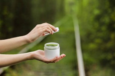 Genç kadın elleri yüz ya da vücut için beyaz bir krem kavanozunu açar. Doğal yeşil arka planda organik doğal cilt ürünleri. Losyon ya da krem paketi. Güzellik kozmetik cilt bakımı