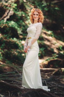 Ormanda, başında çiçekli bir çelenkle güzel, kızıl saçlı bir gelin. Yaz günü açık havada uzun beyaz elbiseli bir kadın. düğün günü.