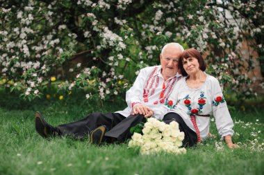 Ukrayna 'nın ulusal işlenmiş tişörtleri içinde çimlerin üzerinde oturan güzel bir çift. Eski bir çiftin evlilik yıldönümü. Büyük bir buket beyaz gül.