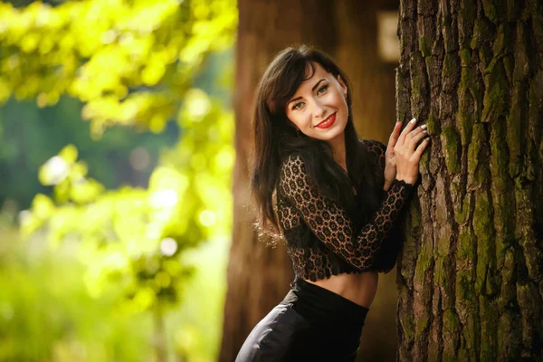 Szexi barna lány csipke blúz és bőr szoknya magas sarkú pózol közel egy fa az erdőben — Stock Fotó