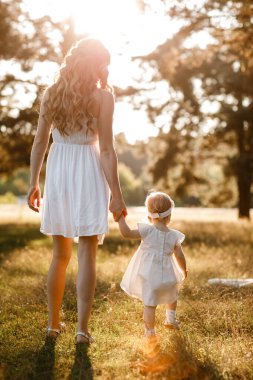 Genç ve güzel anne güneşli parkta küçük kızıyla yürüyor. Açık havada yaz tatili kavramı. Anneler günü, bebekler günü. Birlikte vakit geçirmek. Güneş ışığı. arka plan.