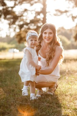 Genç ve güzel anne güneşli parkta küçük kızıyla yürüyor. Açık havada yaz tatili kavramı. Anneler günü, bebekler günü. Birlikte vakit geçirmek. Güneş ışığı