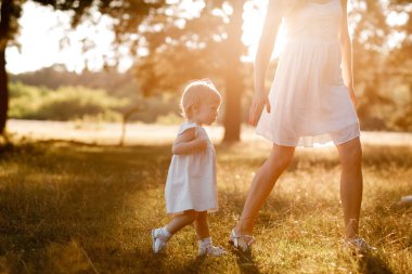 Genç ve güzel anne güneşli parkta küçük kızıyla yürüyor. Açık havada yaz tatili kavramı. Anneler günü, bebekler günü. Birlikte vakit geçirmek. Güneş ışığı