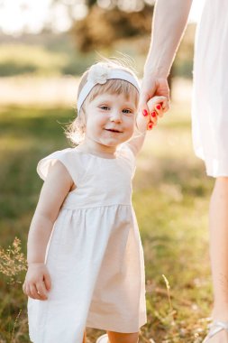 Yeni yürümeye başlayan çocuğa bak. Anne el ele tutuşur kız doğanın tadını çıkarır ve yaz parkında yürür. Genç bir aile tatilde, dışarıda birlikte vakit geçiriyor. Anneler günü, bebekler günü..