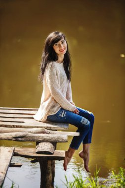 Yaşlı köprüde çıplak ayakla oturan genç bir kadın. Yaz, yeşil orman, akşam göl ve hava dinlenmek, meditasyon yapmak, doğanın seslerinin tadını çıkarmak ve rüya görmek için ortam.