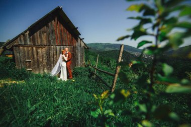 Dağlarda bir düğün çifti. Damat ve gelin kırsal evin yakınında kucaklaşıyorlar. Çiftin arkasındaki dağların güzel manzarası..