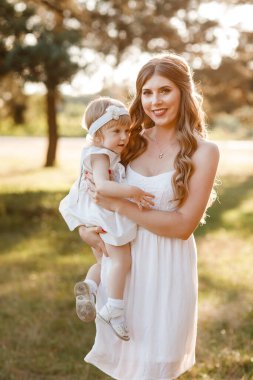 Genç ve güzel bir anne, küçük kızını güneşli parkta tutuyor. Açık havada yaz tatili kavramı. Anneler günü, bebekler günü. Birlikte vakit geçirmek. Güneş ışığı