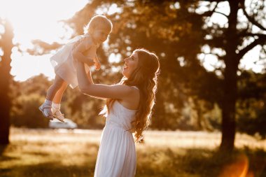 Genç ve güzel anne güneşli parkta küçük kızıyla yürüyor. Açık havada yaz tatili kavramı. Anneler günü, bebekler günü. Birlikte vakit geçirmek. Güneş ışığı