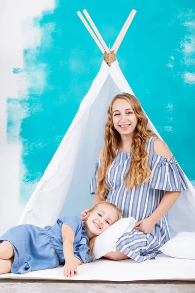 happy pregnant mother and little daughter having fun near the wigwam on the background of a turquoise wall. the girl lies in her mother's lap. mother's day, baby's day. easter.