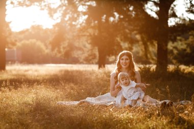 Genç ve güzel anne güneşli parkta küçük kızıyla yürüyor. Açık havada yaz tatili kavramı. Anneler günü, bebekler günü. Birlikte vakit geçirmek. Güneş ışığı