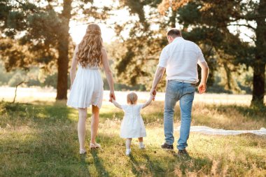 Anne ve babanın el ele tutuşması, kızın doğanın tadını çıkarması ve yaz parkında yürümesi. Genç bir aile tatilde, dışarıda birlikte vakit geçiriyor. Anneler günü, babalar günü, bebekler günü..