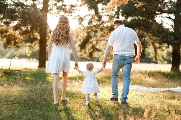 Anne ve babanın el ele tutuşması, kızın doğanın tadını çıkarması ve yaz parkında yürümesi. Genç bir aile tatilde, dışarıda birlikte vakit geçiriyor. Anneler günü, babalar günü, bebekler günü..