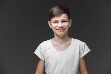 Beyaz tişörtlü yakışıklı çocuk gri boş duvarın önünde poz veriyor. Modaya uygun erkek çocuk portresi. Gülümseyen çocuk poz veriyor, arka planda boş duvar var. Çocuk stili ve moda anlayışı.