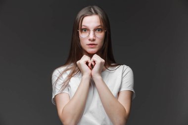 Çekici, heyecanlı, korkmuş, korkmuş genç kız gözlük takıyor, beyaz tişört giyiyor, kahverengi uzun saçlı, gri arka planda izole edilmiş. İnsan duyguları kavramı, yüz ifadesi, satış, reklam