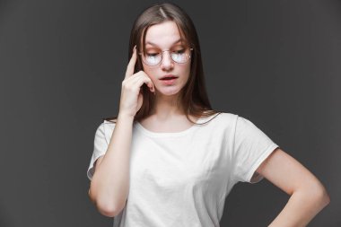 Genç ve güzel bir kadın, boş alana bakıyor. Gözlüklü ve beyaz tişörtlü kız, gri arka planda izole edilmiş..