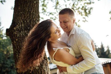 Sevgi dolu genç bir çift yaz güneşli bir günde dışarıda eğleniyor. Erkek ve kadın parkta ağacın yanında sarılıyorlar. Sevgililer Günü.