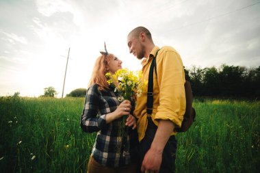 Bir buket kır çiçeği kadının elinde. Sevgi dolu hipster bir çift sahada yürüyor, öpüşüyor ve el ele tutuşuyor, sarılıyor, yazın günbatımında çimlerin üzerinde uzanıyor. Sevgililer Günü.