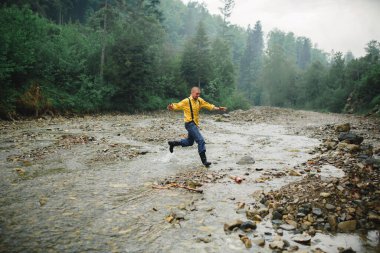 Genç bir sporcu dağ nehrinden aşağı koşar, dağlık bir dağda kayaların üzerinden atlar ve yaz günü yeşil kozalaklı ve yaprak döken bir ormandır.