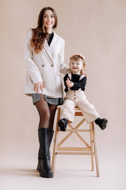 Genç ve güzel bir kız çocuğu olan zarif bir annenin geniş görüşü. Stüdyoda bej arka planda merdivende oturan sevimli, gülümseyen bir çocuk. Anneler günü, bebekler günü. Aile birlikte eğleniyor.