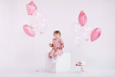 Pembe elbiseli iki yaşındaki küçük kızın doğum günü kutlu olsun. Mumlu ve güllü beyaz pasta. Beyaz renkli balonlar ve beyaz bir duvarda parti için konfeti olan doğum günü süslemeleri. Mutlu yıllar..